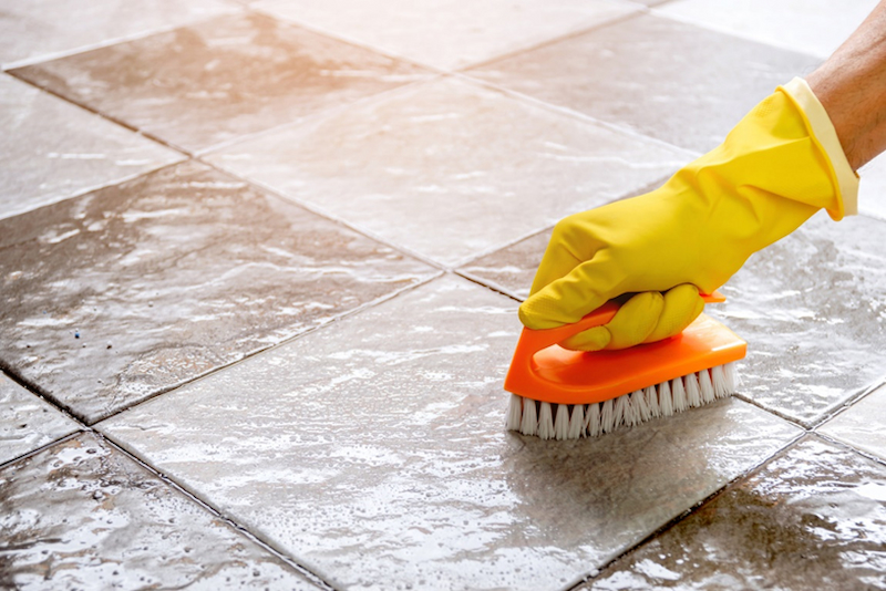 A person cleaning a tile floor with yellow gloves and scrubbing brush